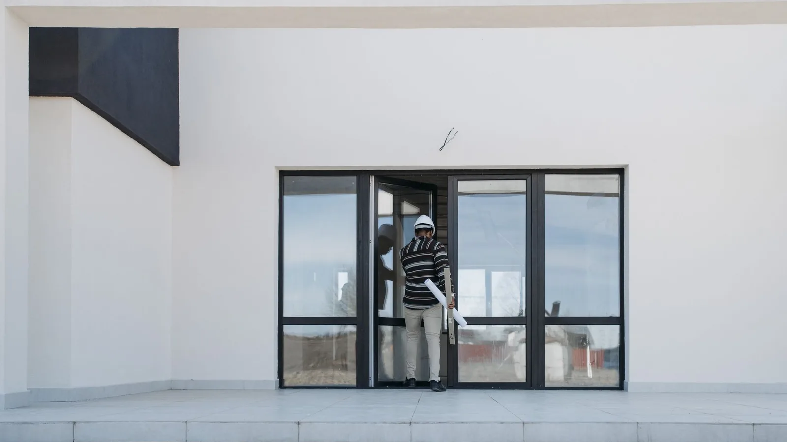Architect wearing helmet and holding blueprints exits a modern glass building