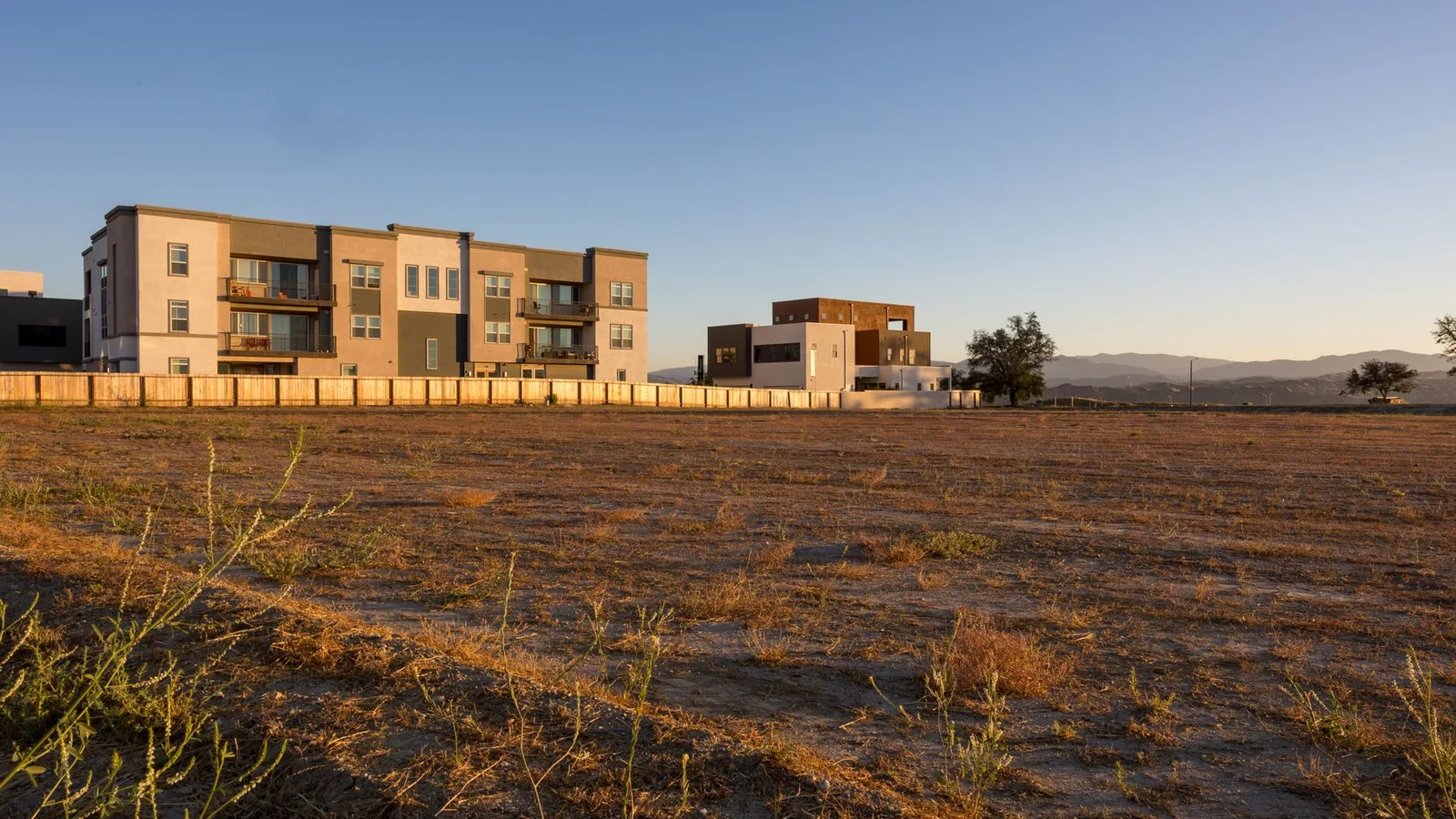 Contemporary apartment blocks with ample open field space, framed by distant mountains under a clear blue sky