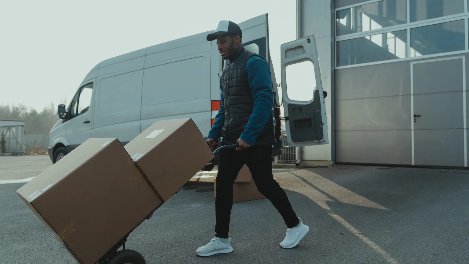 Courier pushing a trolley of boxes past a delivery van at a warehouse loading bay